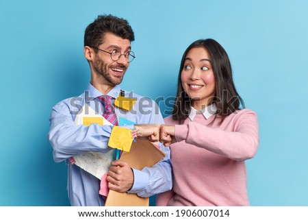 Cheerful mixed race woman and man colleagues celebrate successfully finished work make fist bump pose with paper documents look gladfully at each other isolated over blue background. Partnership