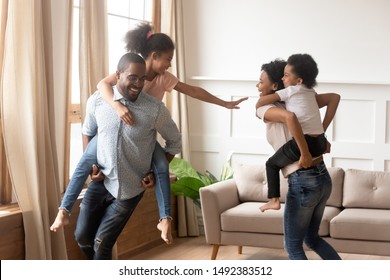 Cheerful mixed-race family playing in living room feels happy move at new house, mother hold piggyback riding son, father carrying daughter people having fun enjoy leisure active game together at home