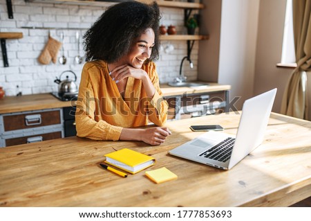 A cheerful mixed-race girl uses laptop for remote work or home leisure while sitting in the kitchen at home. Side view a nice girl with an afro hairstyle looks at screen with a smile