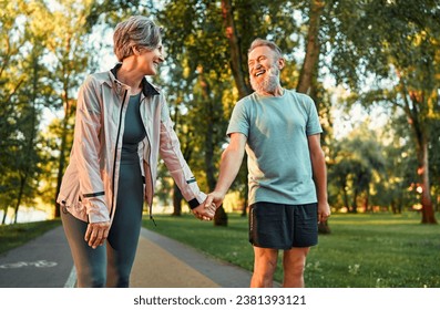 Cheerful modern senior couple outdoors in the park dressed in sportswear walking holding hands and laughing. Morning sun rays fall on smiling man's face.
 - Powered by Shutterstock