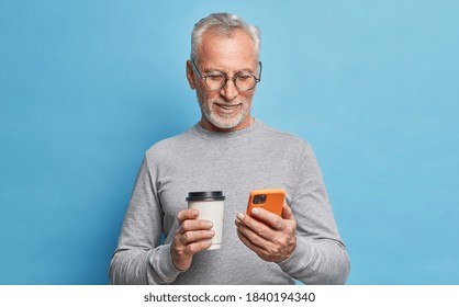 Cheerful modern senior man uses cellphone for communication types text message on phone screen holds paper cup of coffee scrolls internet pages dressed in casual clothes isolated over blue wall