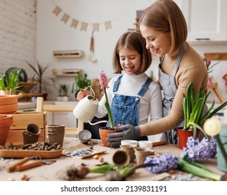 Cheerful Mom In Casual Clothes Smiling And Embracing Kid While Watering Potted Hyacinth Together On Messy Table In Light Kitchen At Home