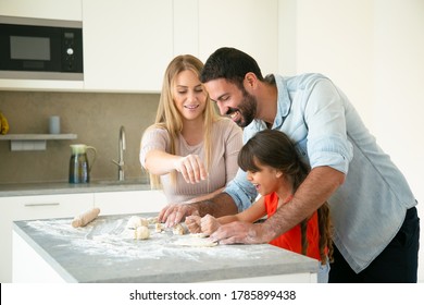 Cheerful Mom And Dad Teaching Daughter To Make Dough On Kitchen Table With Flour Messy. Young Couple And Their Girl Baking Buns Or Pies Together. Family Cooking Concept