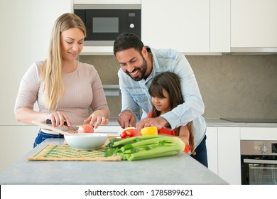 Cheerful mom and dad teaching kid to cook. Young couple and their girl cutting fresh fruits and vegetables for salad at kitchen counter. Healthy nutrition or lifestyle concept - Powered by Shutterstock