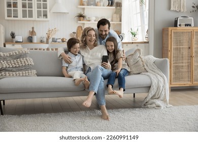 Cheerful mom, dad and two little kids using mobile phone at home together, hugging on sofa, playing online virtual game, taking self video for blog, social media, making family call - Powered by Shutterstock