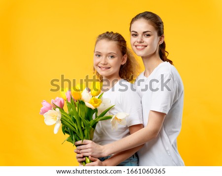 Cheerful Mom and daughter hugs White t-shirts flowers holiday decoration smile yellow background