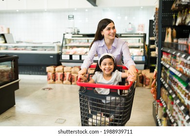 Cheerful mom and her adorable daughter smiling while playing with a shopping cart at a supermarket aisle 