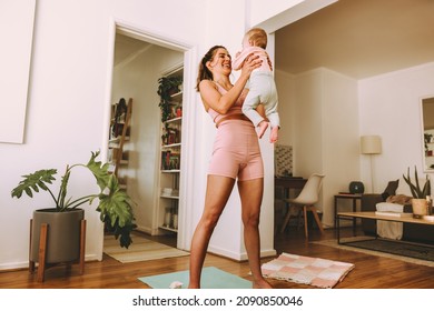 Cheerful Mom Smiling At Her Baby While Lifting Her Up. Happy Young Mom Working Out With Little Baby At Home. New Mom Bonding With Her Baby During Her Post-natal Fitness Routine.