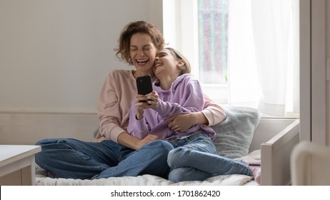 Cheerful mom and teen daughter laughing using smartphone app at home sitting on bed. Happy positive family adult parent having fun watching funny videos enjoying lifestyle relaxing with teenage girl.