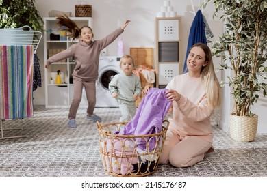 A Cheerful Mom With Two Daughters Organizes Clothes In The Laundry Room,bathroom. The Woman Is Kneeling By The Wicker Basket And Sorting Dirty Things, While The Girls Are Running Around, Playing.