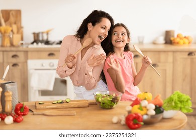 Cheerful mommy and daughter kid using spoons as microphones, singing while making fresh salad together, standing near a table with organic vegetables in modern kitchen, enjoying family cooking fun - Powered by Shutterstock