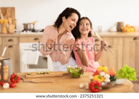 Cheerful mommy and daughter kid using spoons as microphones, singing while making fresh salad together, standing near a table with organic vegetables in modern kitchen, enjoying family cooking fun