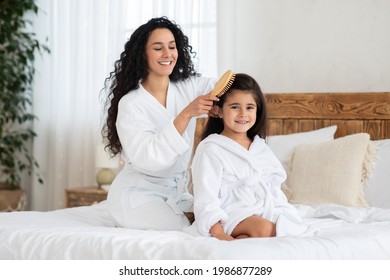 Cheerful mother brushing her cute little daughter long curly hair while sitting together on bed at home. Beautiful young woman parent taking care about her child hair, combing with wooden brush - Powered by Shutterstock