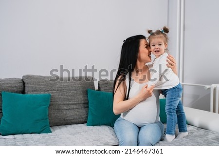 Cheerful mother and daughter are on the sofa.Pregnant mom's face turned to her daughter's face and little girl with ponytails looks at camera and smiles.A cute child stands on couch next to her mother