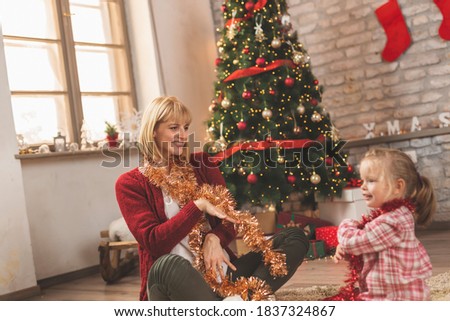 Cheerful mother and daughter sitting by nicely decorated Christmas tree, having fun while playing at home on Christmas day