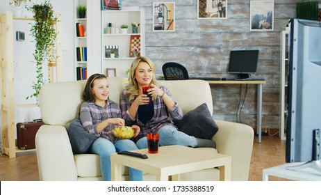 Cheerful Mother And Daughter Sitting On The Couch In Living Room Watching Tv, Eating Chips And Drinking Soda.