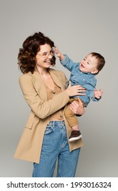 Cheerful Mother In Glasses And Blazer Holding In Arms Happy Baby Boy Isolated On Grey