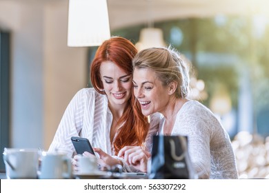 Cheerful mother and her daughter sit in a cafe, they laugh while watching pictures on a phone screen. Shot with flare