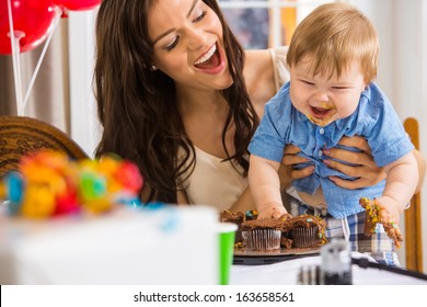 Cheerful Mother Holding Baby Boy With Messy Hands Covered With Cake Icing At Birthday Party