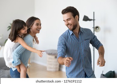 Cheerful mother laughing piggybacking little daughter playing at home, happy mom carrying child girl on back having fun catching dad, joyful parents and kid running enjoy funny game in living room