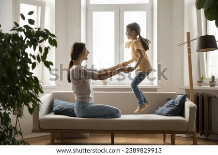 Cheerful mother play with little cute daughter on couch, mom holding hands while joyful girl jumping on soft sofa. Happy family spend time at home, enjoy warm relationship, have fun. Motherhood, love