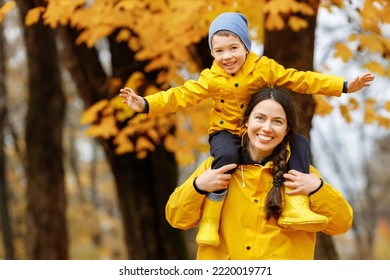 Cheerful Mother In Yellow Raincoat Holding Her Son On Shoulder Having Fun In The Autumn Park. Concept Of Happy Family