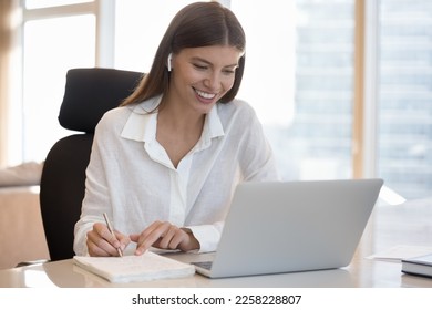 Cheerful motivated pretty student woman with wireless earbud in ear talking on video call at computer, writing notes, watching learning webinar, using laptop, speaking at online conference - Powered by Shutterstock