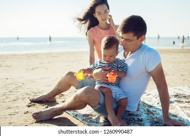 Cheerful Multi Ethnic Family Have A Rest On A Sea Shore. Young Family, Man Woman And Little Son Enjoy Picnic, Summer Outdoor, Good Weather