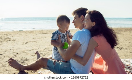 Cheerful Multi Ethnic Family Have A Rest On A Sea Shore. Young Family, Man Woman And Little Son Enjoy Picnic, Summer Outdoor, Good Weather