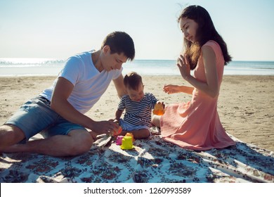 Cheerful Multi Ethnic Family Have A Rest On A Sea Shore. Young Family, Man Woman And Little Son Enjoy Picnic, Summer Outdoor, Good Weather