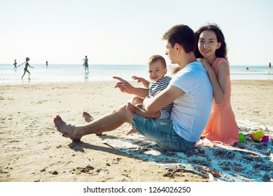 Cheerful Multi Ethnic Family Have A Rest On A Sea Shore. Young Family, Man Woman And Little Son Enjoy Picnic, Summer Outdoor, Good Weather