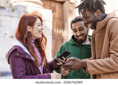 Cheerful multicultural friends looking at smartphone together in urban setting. Young diverse group sharing funny content on social media, representing modern friendship and technology use everyday - Powered by Shutterstock