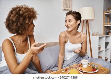 cheerful multicultural lesbian couple in pajamas gesturing and chatting near breakfast in bed - Powered by Shutterstock