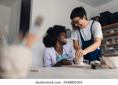 Cheerful multicultural pottery course attendee sitting on a class and making earthenware with her mentor while smiling at her. An african american woman is smiling at her tutor at occupational therapy - Powered by Shutterstock