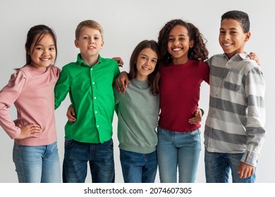 Cheerful Multicultural Preteen Kids Standing Hugging And Smiling To Camera Posing Together Over Gray Studio Background. Shot Of Mixed School Boys And Girls Group. Friendship Concept