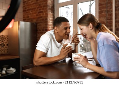 Cheerful Multiethnic Couple Eating Noodles During Making Eat Fast Food Video Challenge On Smartphone. Video Blogging. Man And Woman Of Generation Z Creating Content For Social Networks At Home Kitchen