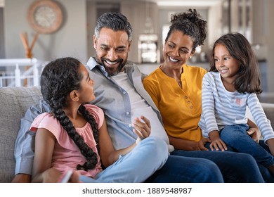 Cheerful Multiethnic Family At Home Sitting On Sofa Listening To Daughter. Happy Couple With Two Girls Relaxing At Home Together. Smiling Indian Parents Talking To Their Daughter In The Living Room.