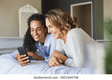 Cheerful multiethnic female friends doing online shopping while lying on bed and using smartphone. Two young women watching funny videos on mobile phone. Lesbian couple using cellphone while relaxing. - Powered by Shutterstock