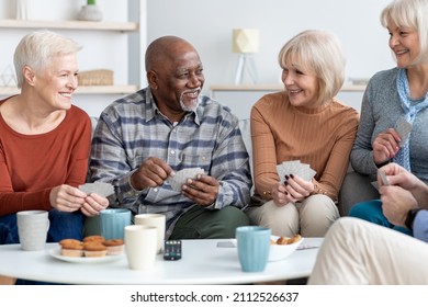 Cheerful Multiethnic Group Of Senior Friends Men And Women In Casual Sitting On Couch, Having Conversation And Playing Cards While Drinking Tea With Cookies, Having Party At Home