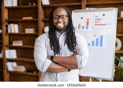 Cheerful multiethnic man with a dreadlocks is online tutor, teacher, a smiling African-American business coach stands in confident pose with folded arms with a flipchart on background, looks at camera - Powered by Shutterstock