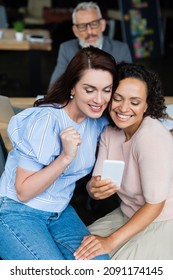 Cheerful Multiethnic Same Sex Couple Looking At Mobile Phone Near Middle Aged Realtor  On Blurred Background