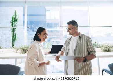 Cheerful multiracial colleagues interacting work details while Asian female looking at screen of tablet in hands of male and standing together at table with laptop against bright sunlight - Powered by Shutterstock