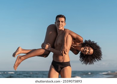 Cheerful multiracial couple enjoying sunset at beach. Young man carrying laughing African American woman with natural curly hair, capturing authentic summer relationship moment. - Powered by Shutterstock