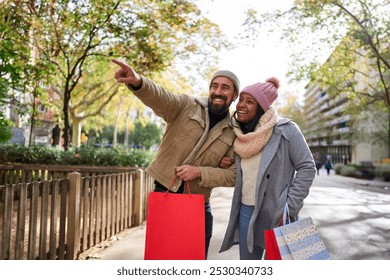 Cheerful multiracial couple walking in the city doing christmas shoppings together, holding gift bags, pointing at things and smiling, having a good time outdoors in winter season. - Powered by Shutterstock