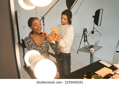 Cheerful multiracial girl holding smartphone and making selfie while make up artist applying make up