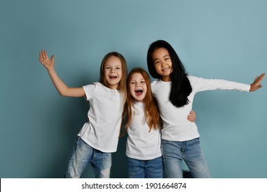 Cheerful multiracial girls friends in jeans and white t-shirts and longsleeves shirt have fun together laughing playing hugging each other over blue background. Happy childhood, stylish look
