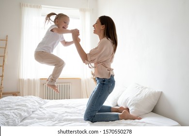 Cheerful mum babysitter play with cute active small kid girl jump on bed, happy carefree mother and little child daughter holding hands laughing having fun feel joy at home in modern bedroom interior