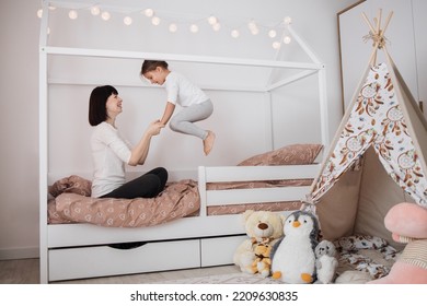 Cheerful Mum Babysitter Play With Cute Active Small Kid Girl Jump On Bed, Happy Carefree Mother And Little Child Daughter Holding Hands Laughing Having Fun Feel Joy At Home In Modern Bedroom Interior