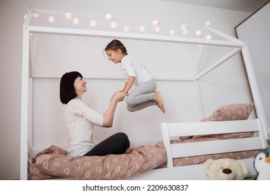 Cheerful Mum Babysitter Play With Cute Active Small Kid Girl Jump On Bed, Happy Carefree Mother And Little Child Daughter Holding Hands Laughing Having Fun Feel Joy At Home In Modern Bedroom Interior