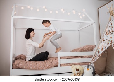 Cheerful Mum Babysitter Play With Cute Active Small Kid Girl Jump On Bed, Happy Carefree Mother And Little Child Daughter Holding Hands Laughing Having Fun Feel Joy At Home In Modern Bedroom Interior